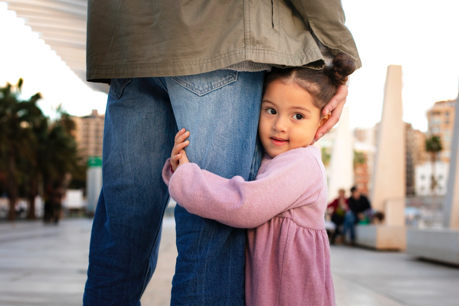 Little girl wearing pink hugs the leg of an adult wearing blue jeans. 