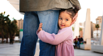 Little girl wearing pink hugs the leg of an adult wearing blue jeans. 