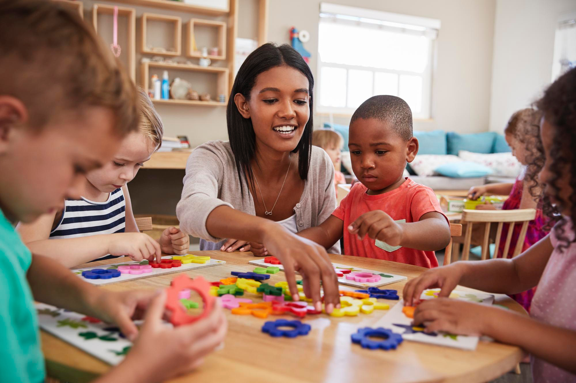 Women sitting at a little table in a preschool classroom setting with 4 little kids, all watching her showing them something with the colorful blocks on the table. 