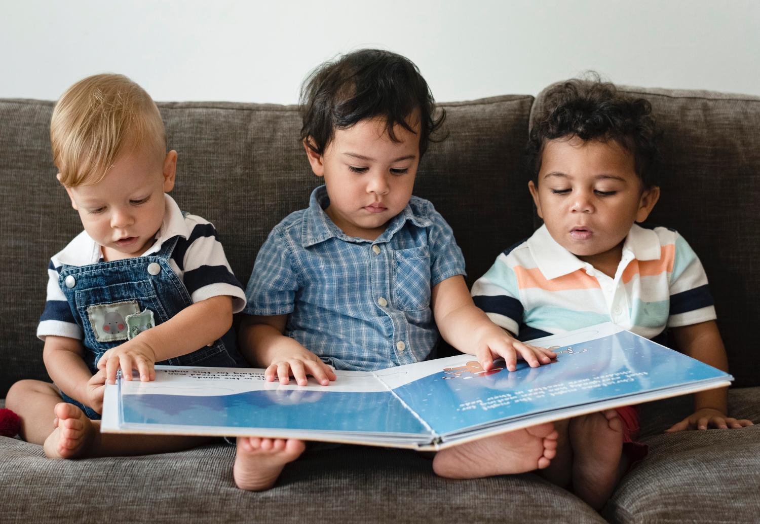 Three toddlers sitting on a couch all looking at the same children's book.