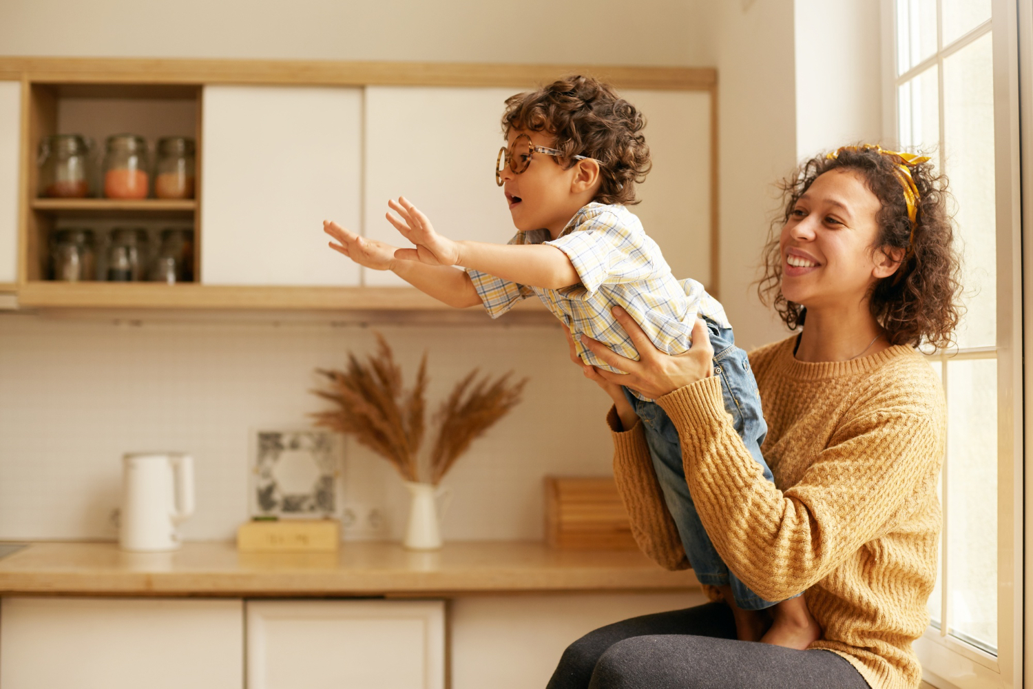 Adult female holds little toddler boy wearing glasses in her lap while he leans forward, both smiling in an indoor home setting. 