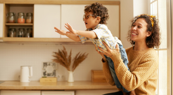 Adult female holds little toddler boy wearing glasses in her lap while he leans forward, both smiling in an indoor home setting. 