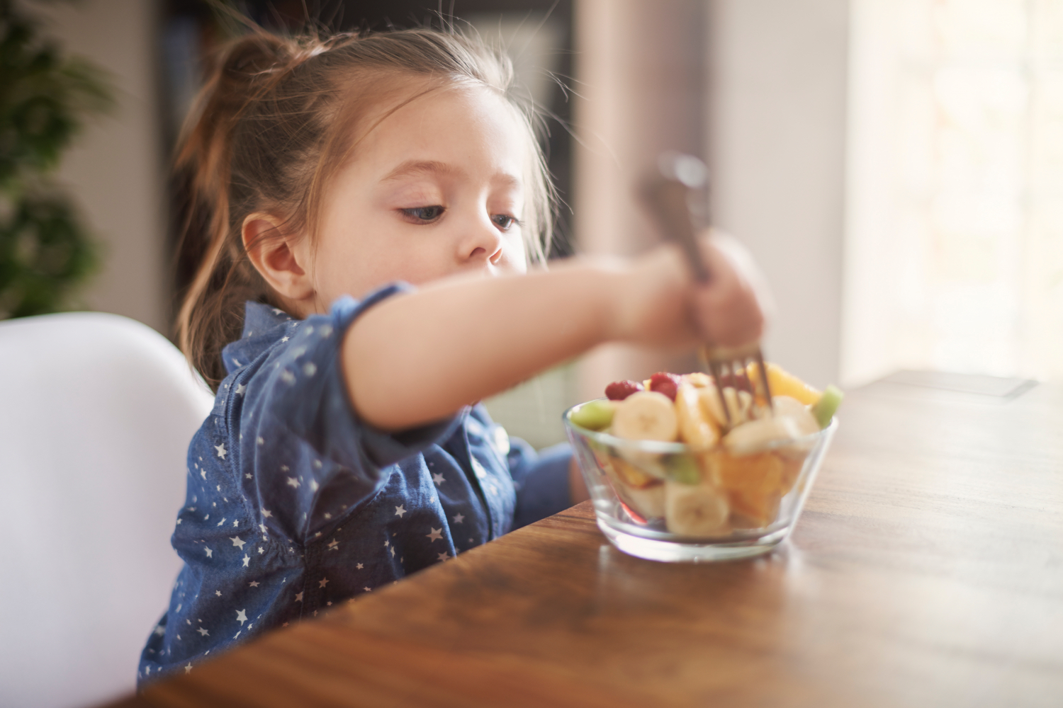 Little girl sitting at table, eating fruit bowl with a fork. 
