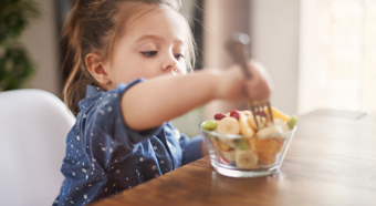 Little girl sitting at table, eating fruit bowl with a fork. 