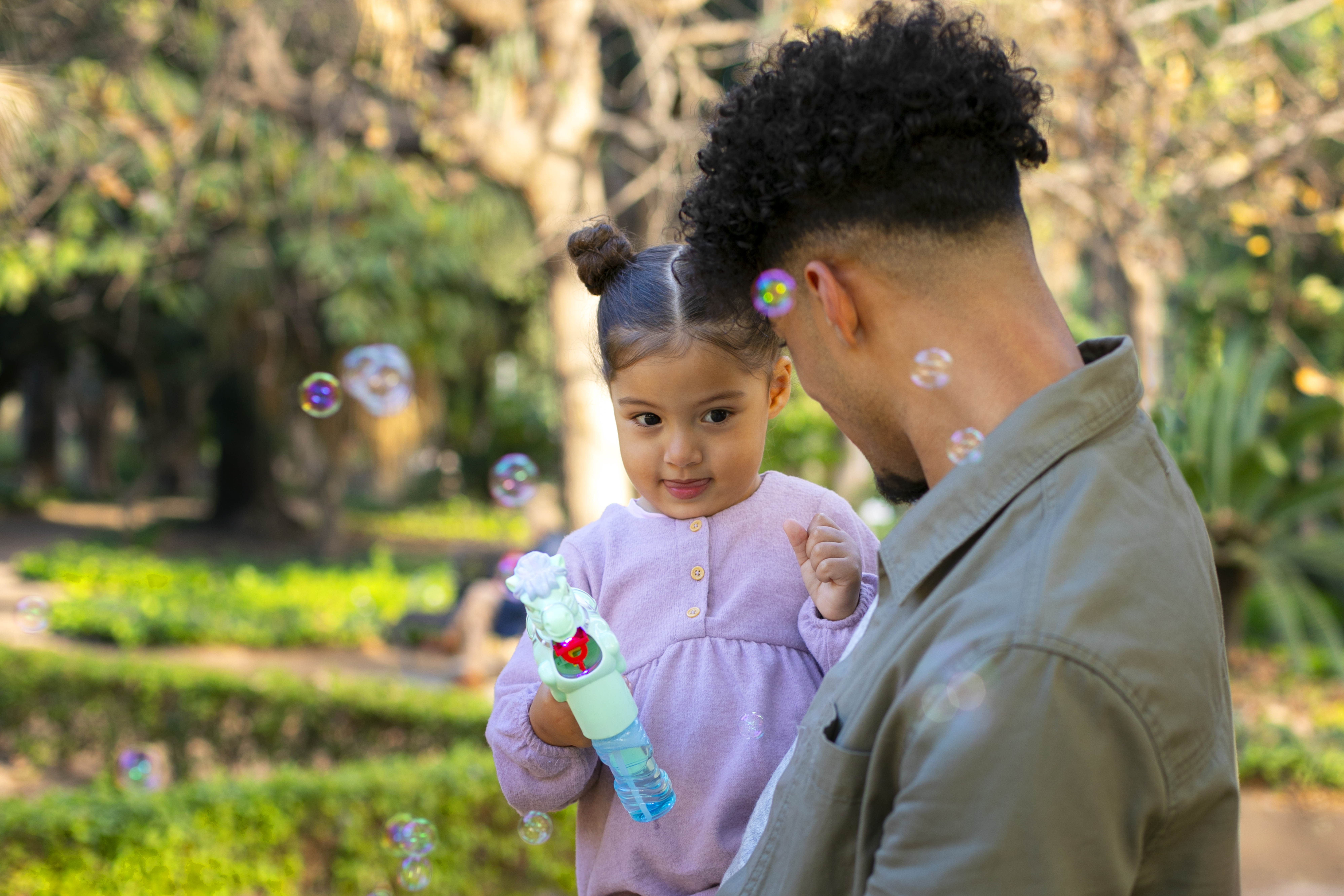 Young girl wearing a purple shirt, holding a bubble blower smiles at adult male who is holding her. In an outdoor green park setting. 