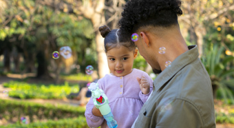 Young girl wearing a purple shirt, holding a bubble blower smiles at adult male who is holding her. In an outdoor green park setting. 