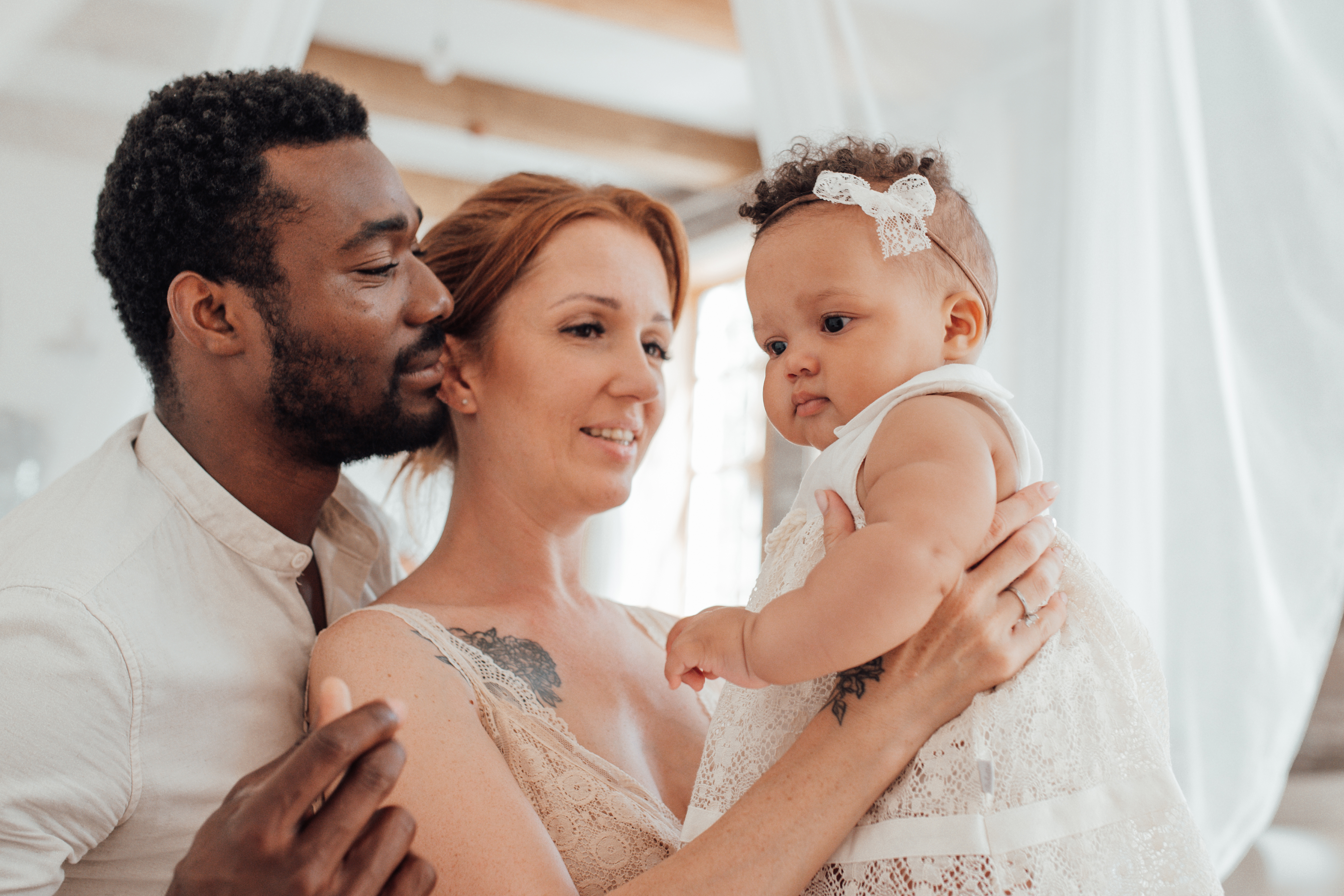 Adult male and female smile and look at baby that the female is holding in front of her. Baby is dressed in a white dress. Indoor bright setting. 