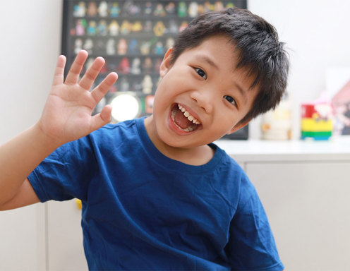 A young boy with light brown skin and brown hair wearing a blue tshirt is smiling and waving to the camera.