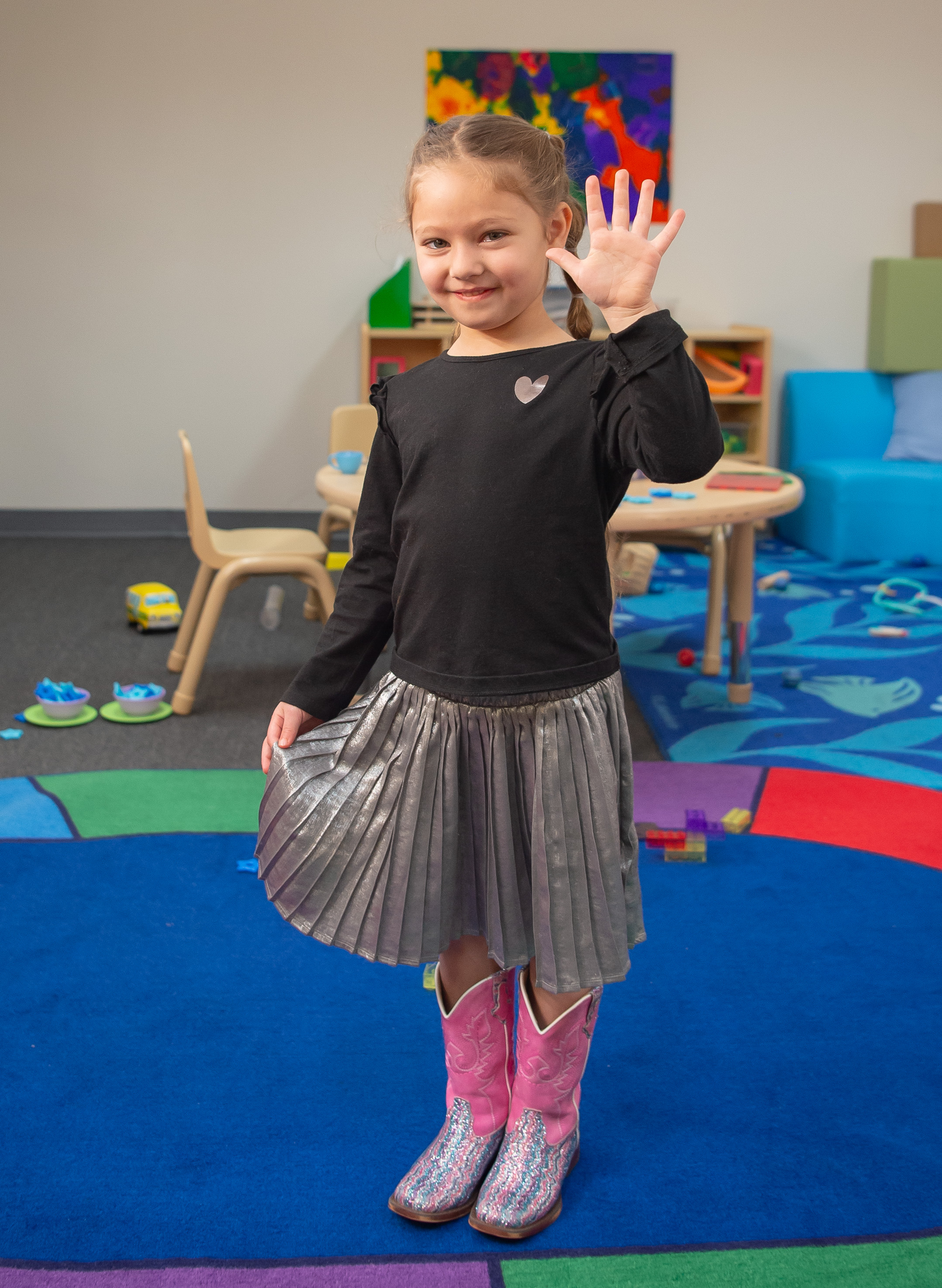 Little girl in colorful classroom setting, wearing pink cowboy boot and putting her hand in the air with five fingers up, smiling at camera. 