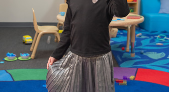 Little girl in colorful classroom setting, wearing pink cowboy boot and putting her hand in the air with five fingers up, smiling at camera. 