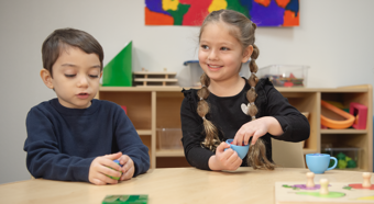 Two toddlers sitting at a table in a classroom setting, smiling playing with toys on the table. 