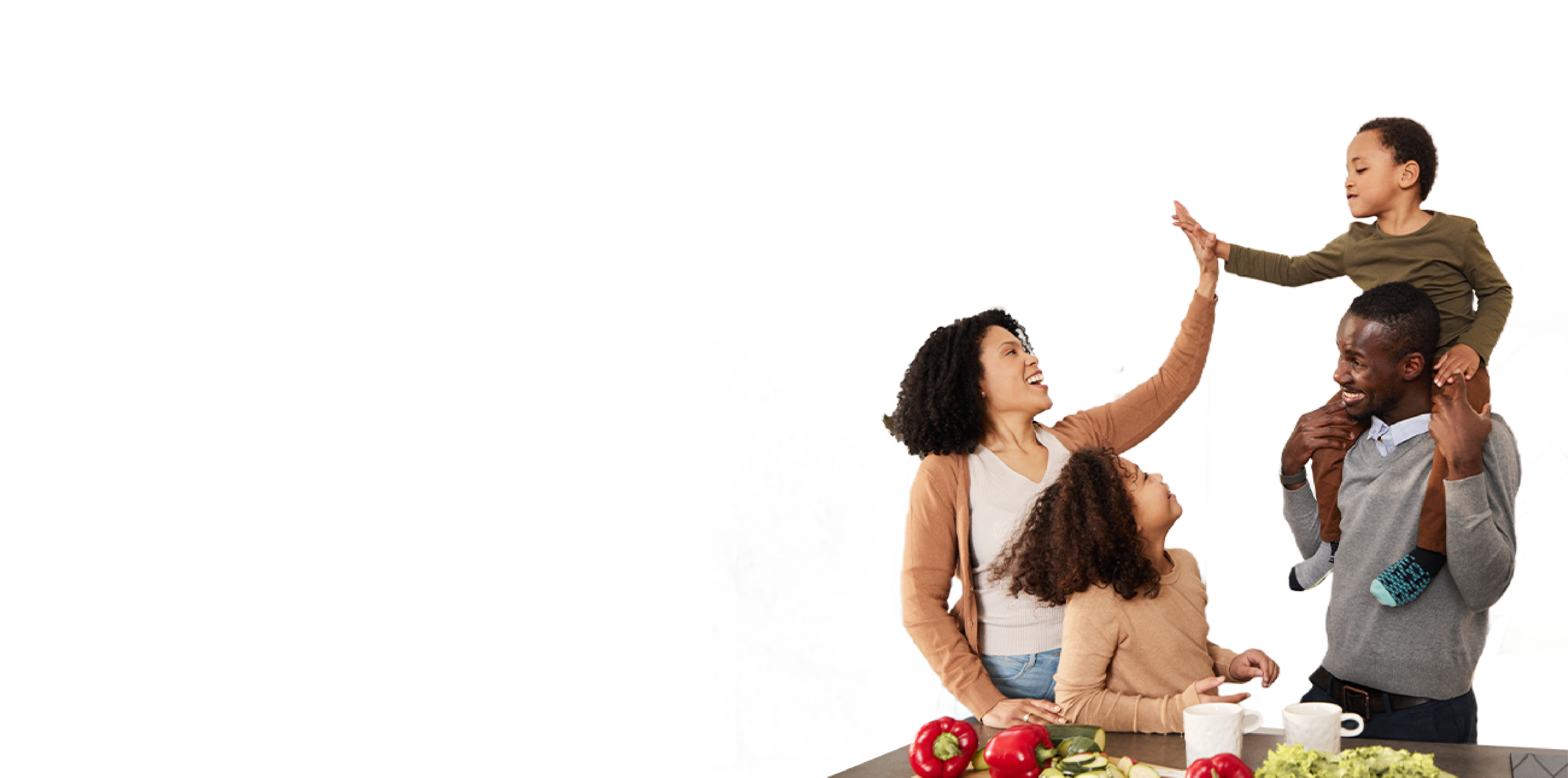 Appearing to be a family unit, a female adult is giving a high five to a young boy who is on the shoulders of the adult male. A young girl is standing beside the adult female smiling up at the boy and male adult. There is a variety of fruits and vegetables on a table beside them.