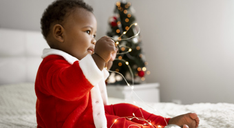 Baby boy dressed up as santa, sitting on bed with Christmas tree in back.