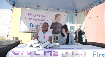 Two young women sitting at a First 5 Nevada booth wave to the camera while smiling, white banners with purple text on the table and behind them. 