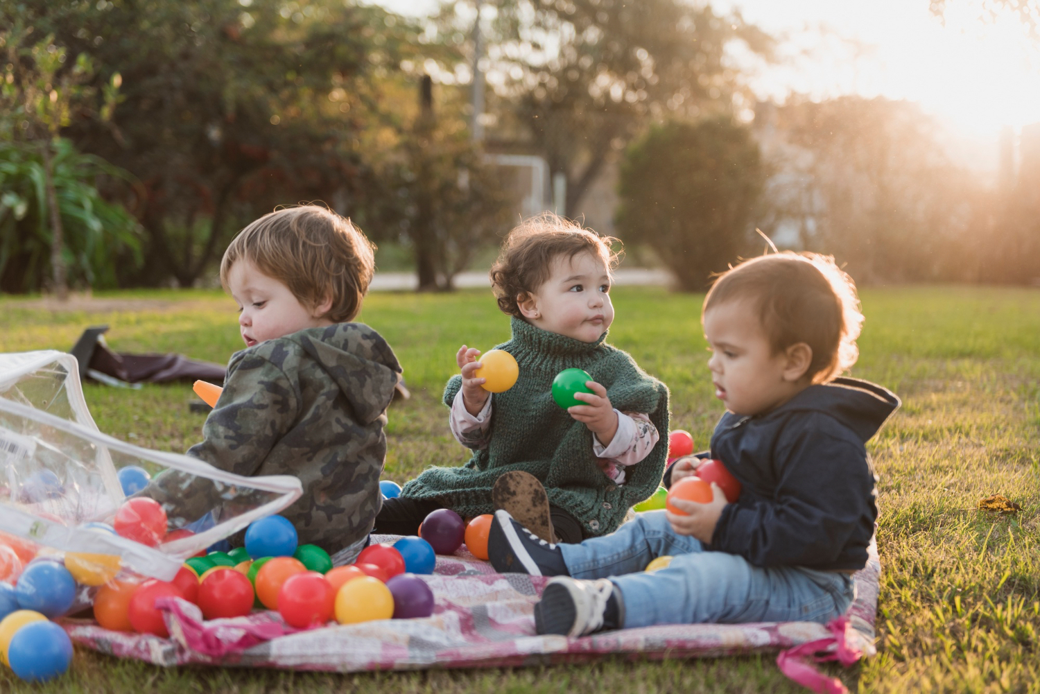 Three toddlers sitting outside in the grass, playing with colorful rainbow toys. 