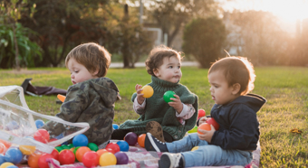 Three toddlers sitting outside in the grass, playing with colorful rainbow toys. 