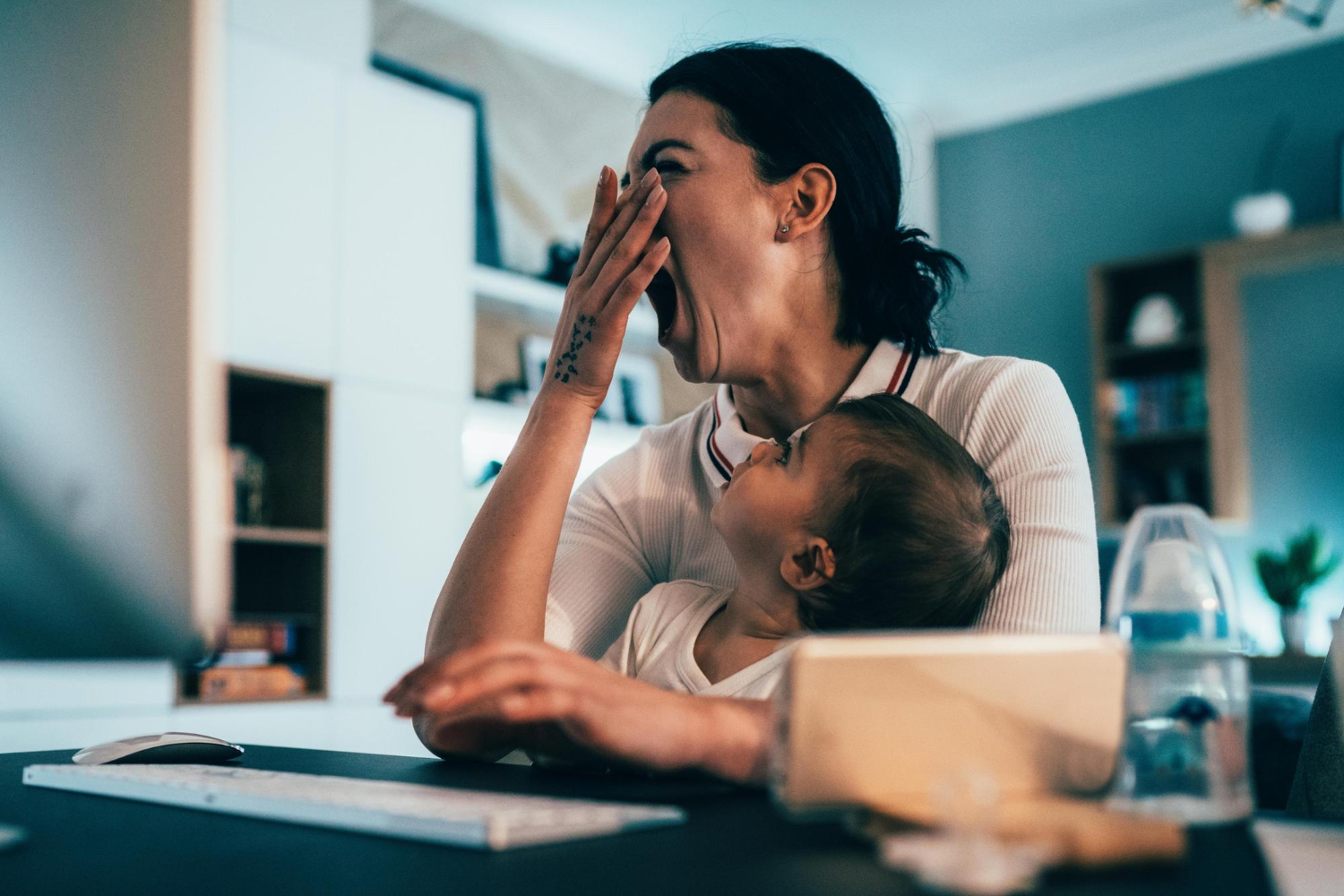 Adult female yawning, with toddler in her lap, sitting at a desk in a home setting. 