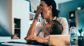 Adult female yawning, with toddler in her lap, sitting at a desk in a home setting. 