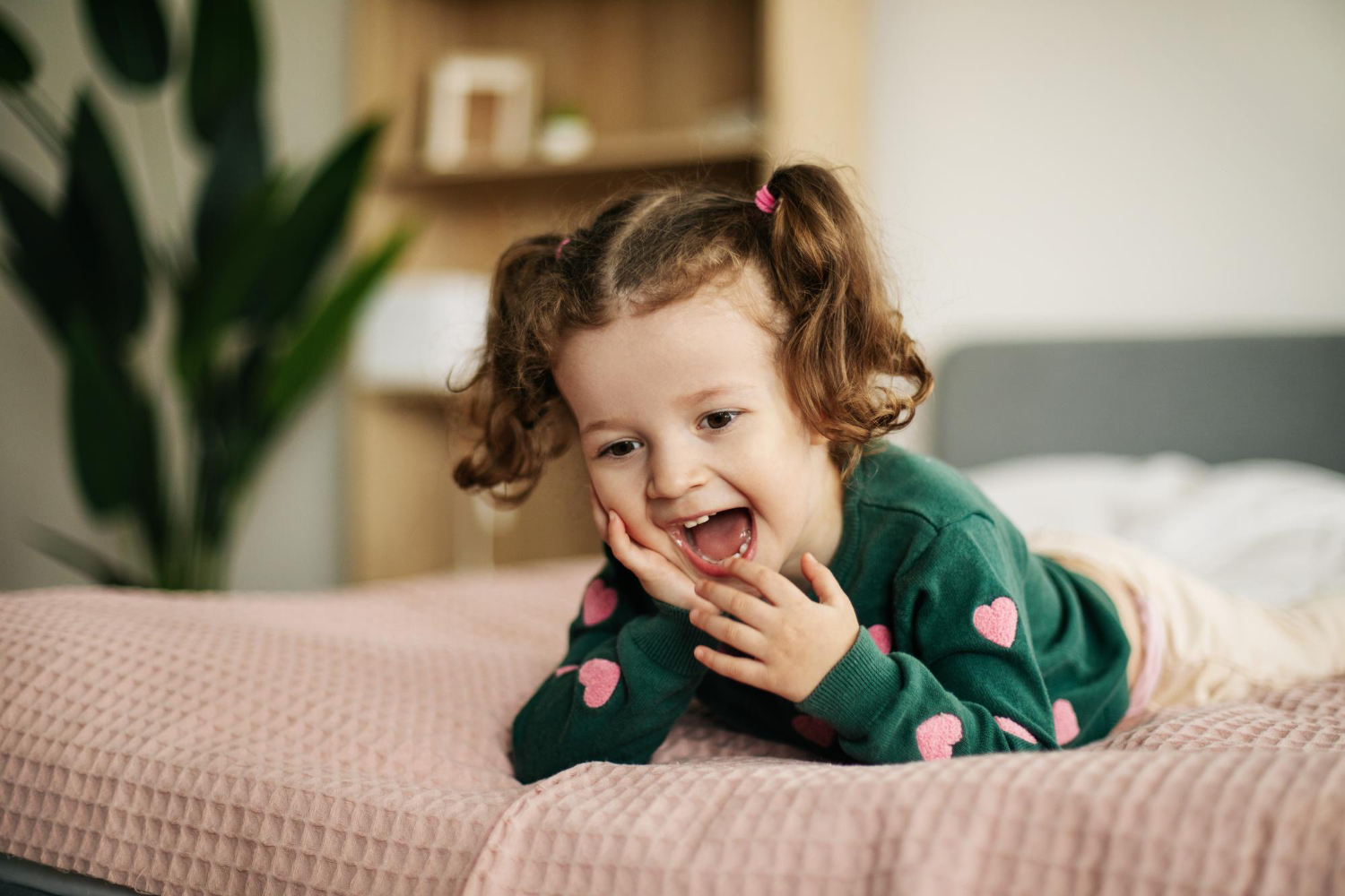 Toddler girl with brown hair smiles while laying belly first on a pink comforter on a bed. 