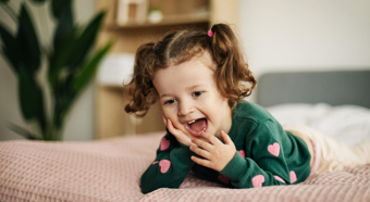 Toddler girl with brown hair smiles while laying belly first on a pink comforter on a bed. 