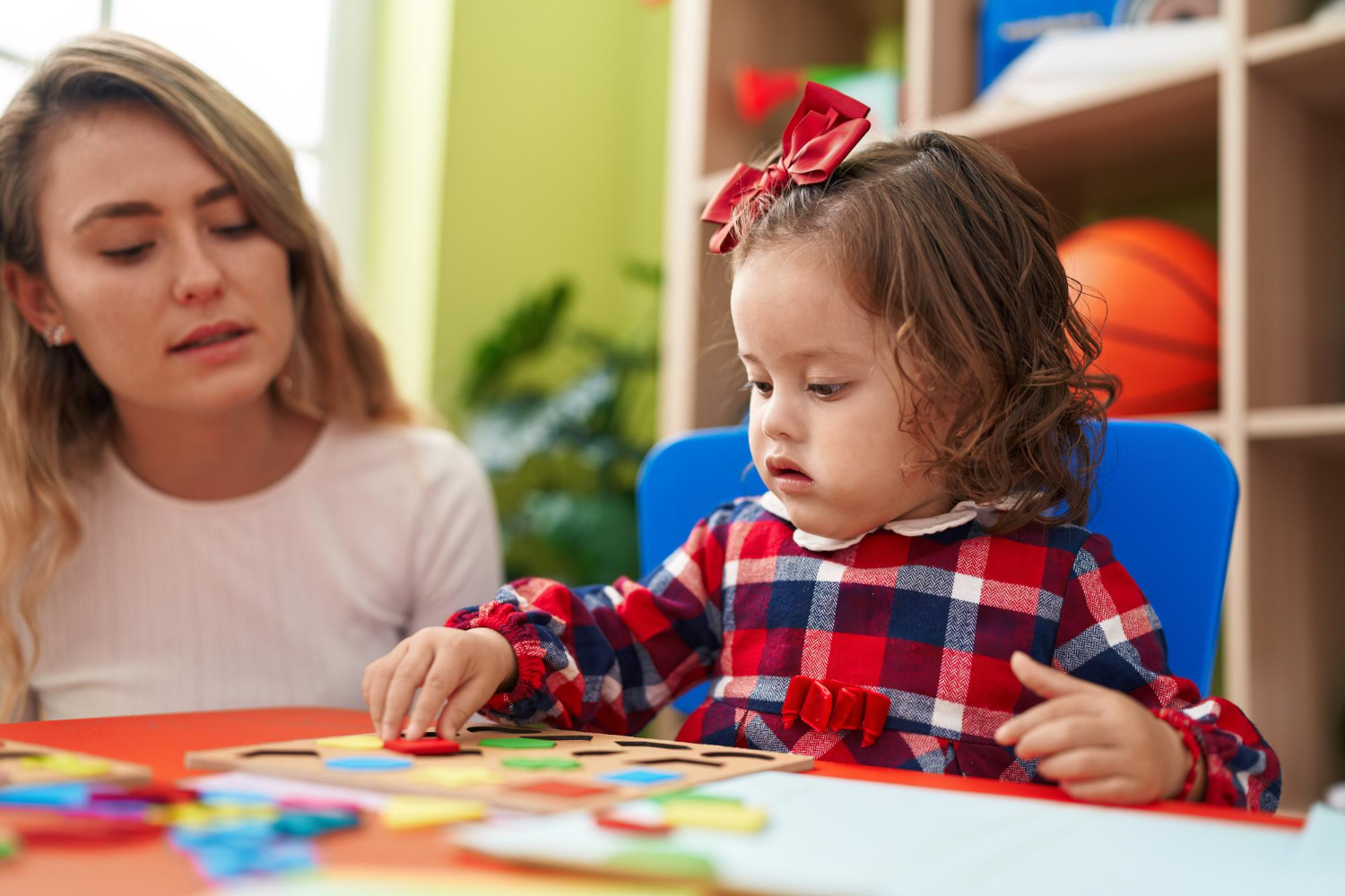 Little girl wearing red plaid shirt plays with a puzzle while adult female sits next to her.