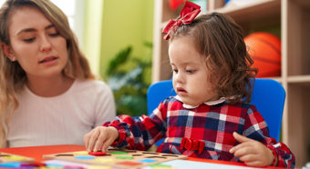 Little girl wearing red plaid shirt plays with a puzzle while adult female sits next to her.