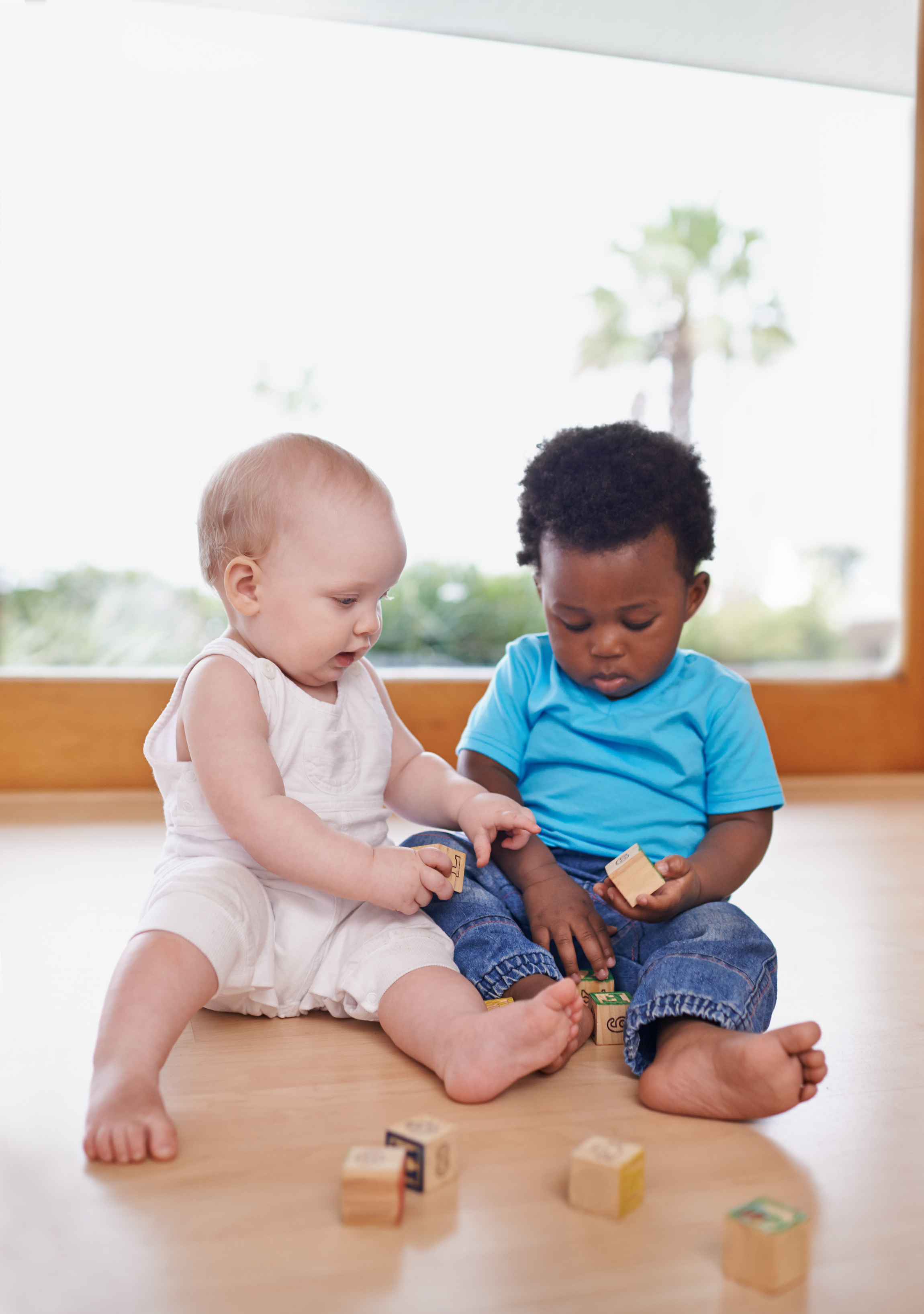 Two toddlers sit on the floor of a home setting while playing with toy blocks. 