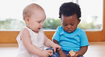 Two toddlers sit on the floor of a home setting while playing with toy blocks. 