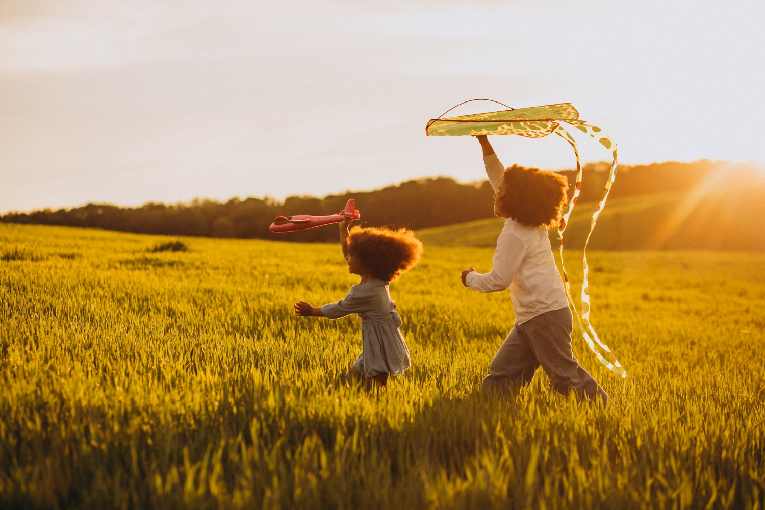 Two young children playing with kites, while running through a grassy field during sunset time. 