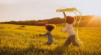 Two young children playing with kites, while running through a grassy field during sunset time. 