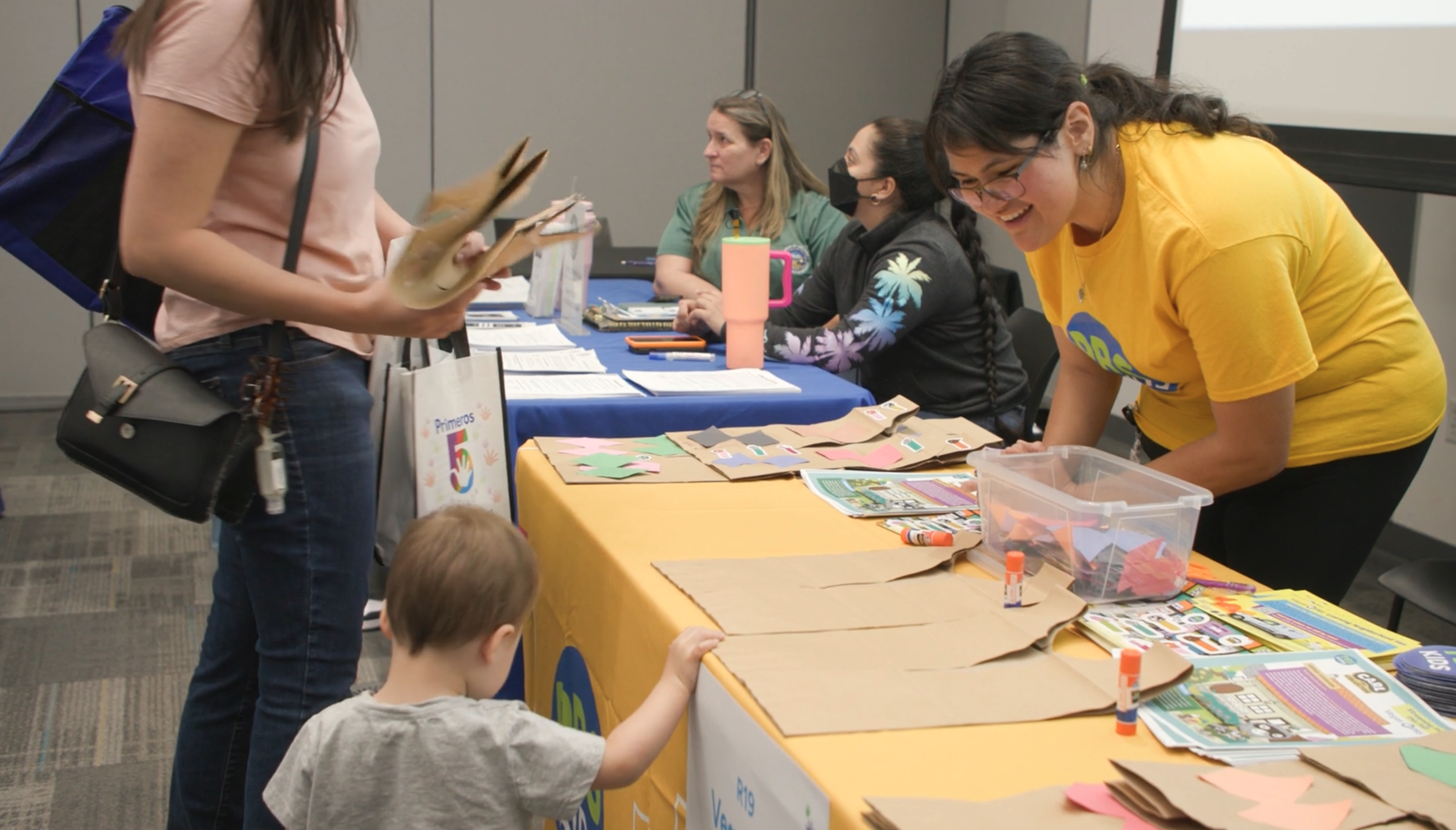 Lady wearing bright yellow shirt, working a table at a community event, smiles at a little toddler and adult female stopping at her table. 