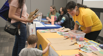 Lady wearing bright yellow shirt, working a table at a community event, smiles at a little toddler and adult female stopping at her table. 