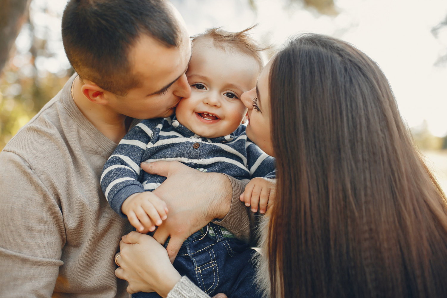 Little toddler being held between two adults who are kissing both cheeks of the smiling toddler. 