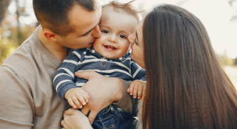 Little toddler being held between two adults who are kissing both cheeks of the smiling toddler. 