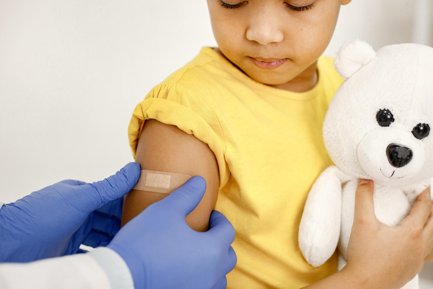 Toddler girl in doctor office setting getting a band aid put on her arm by a doctor, she is holding a stuffed animal.