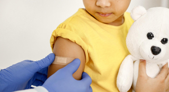 Toddler girl in doctor office setting getting a band aid put on her arm by a doctor, she is holding a stuffed animal.