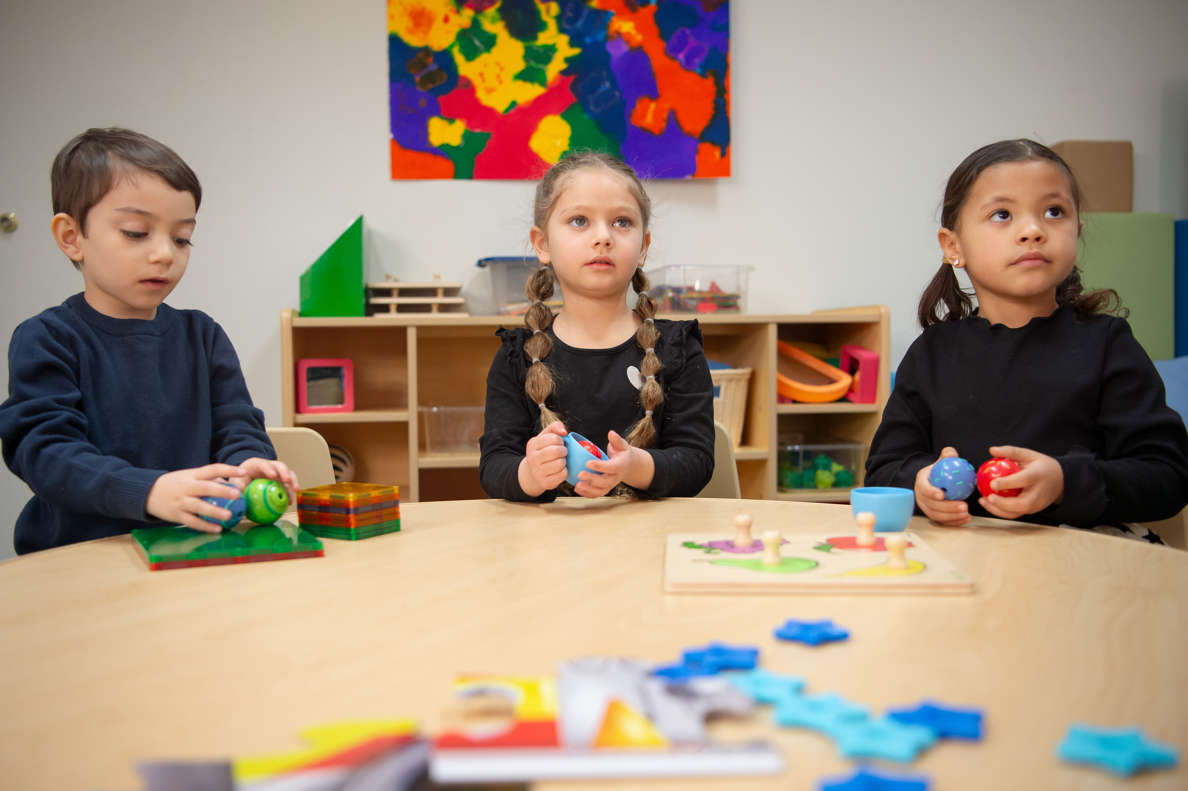 Three toddlers sitting in a colorful classroom setting around a table, with toys on it. 