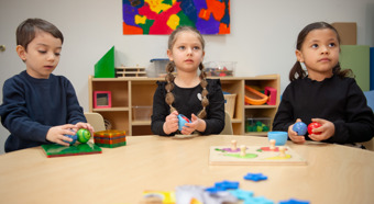 Three toddlers sitting in a colorful classroom setting around a table, with toys on it. 