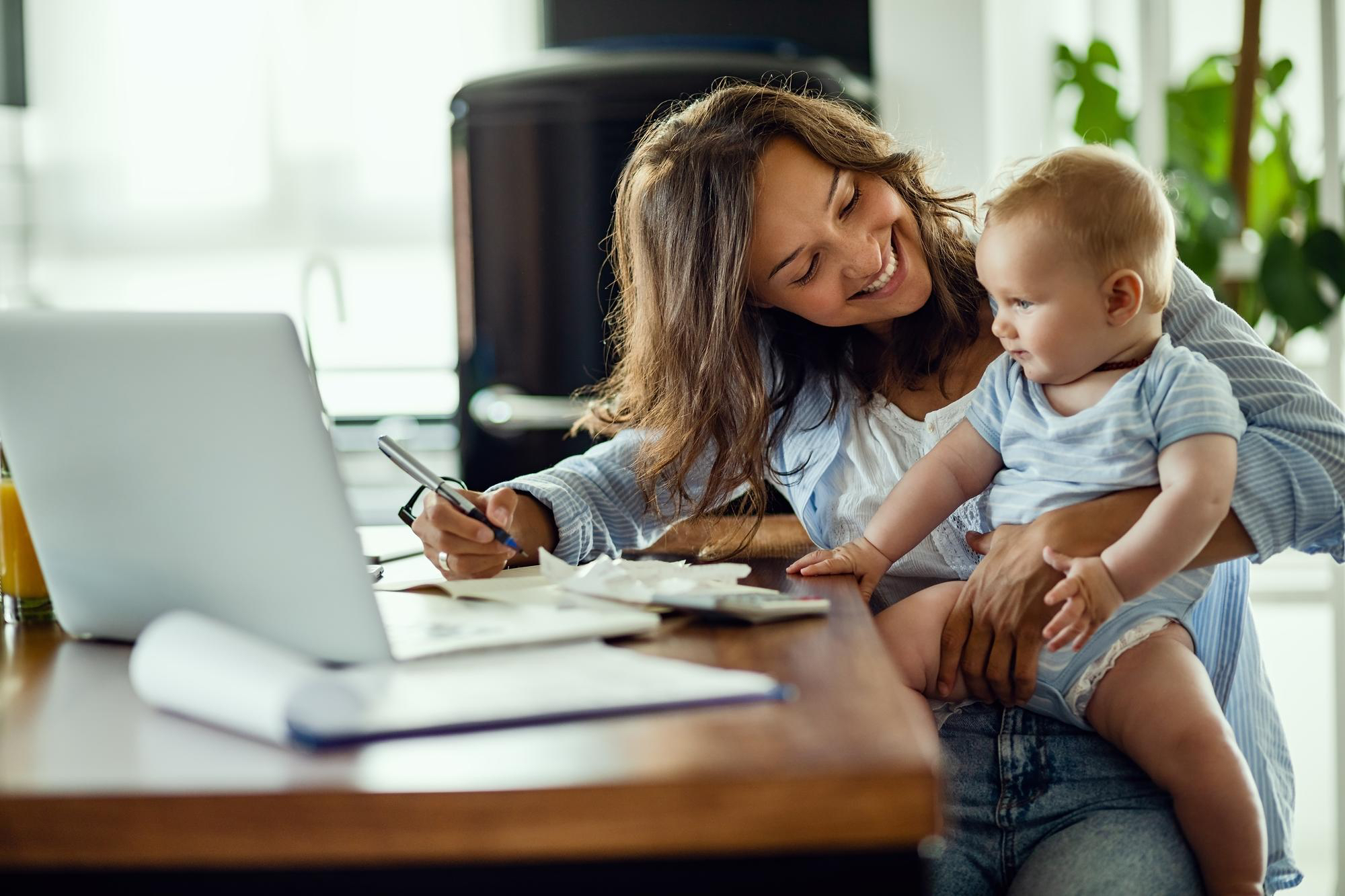 Adult female smiles and toddler in her lap, while at a desk working on her computer. 