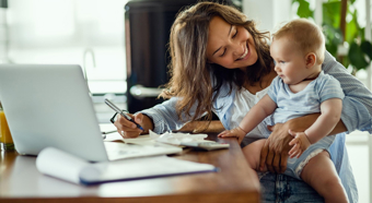 Adult female smiles and toddler in her lap, while at a desk working on her computer. 