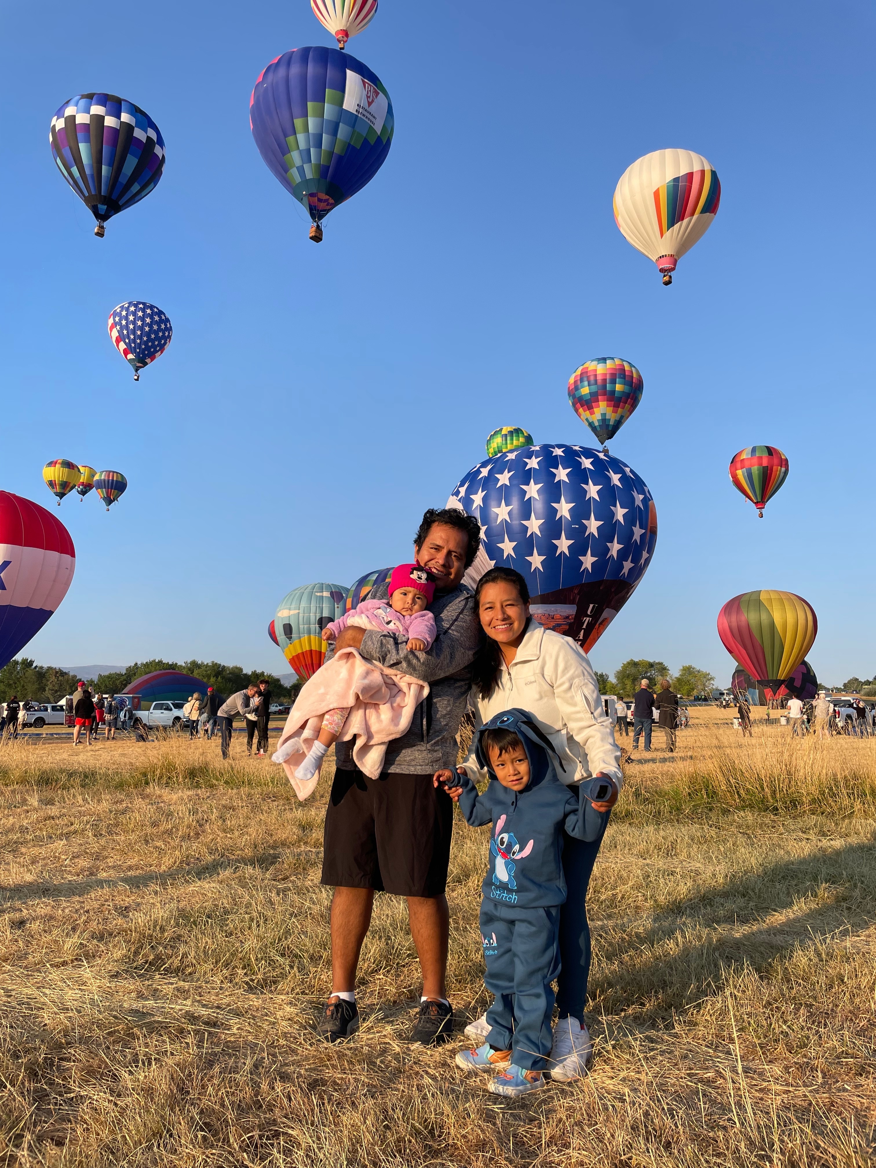 Angela Oscanoa standing with her family in a hot air ballon field. Multiple hot air balloons in the air behind them. 