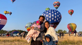 Angela Oscanoa standing with her family in a hot air ballon field. Multiple hot air balloons in the air behind them. 