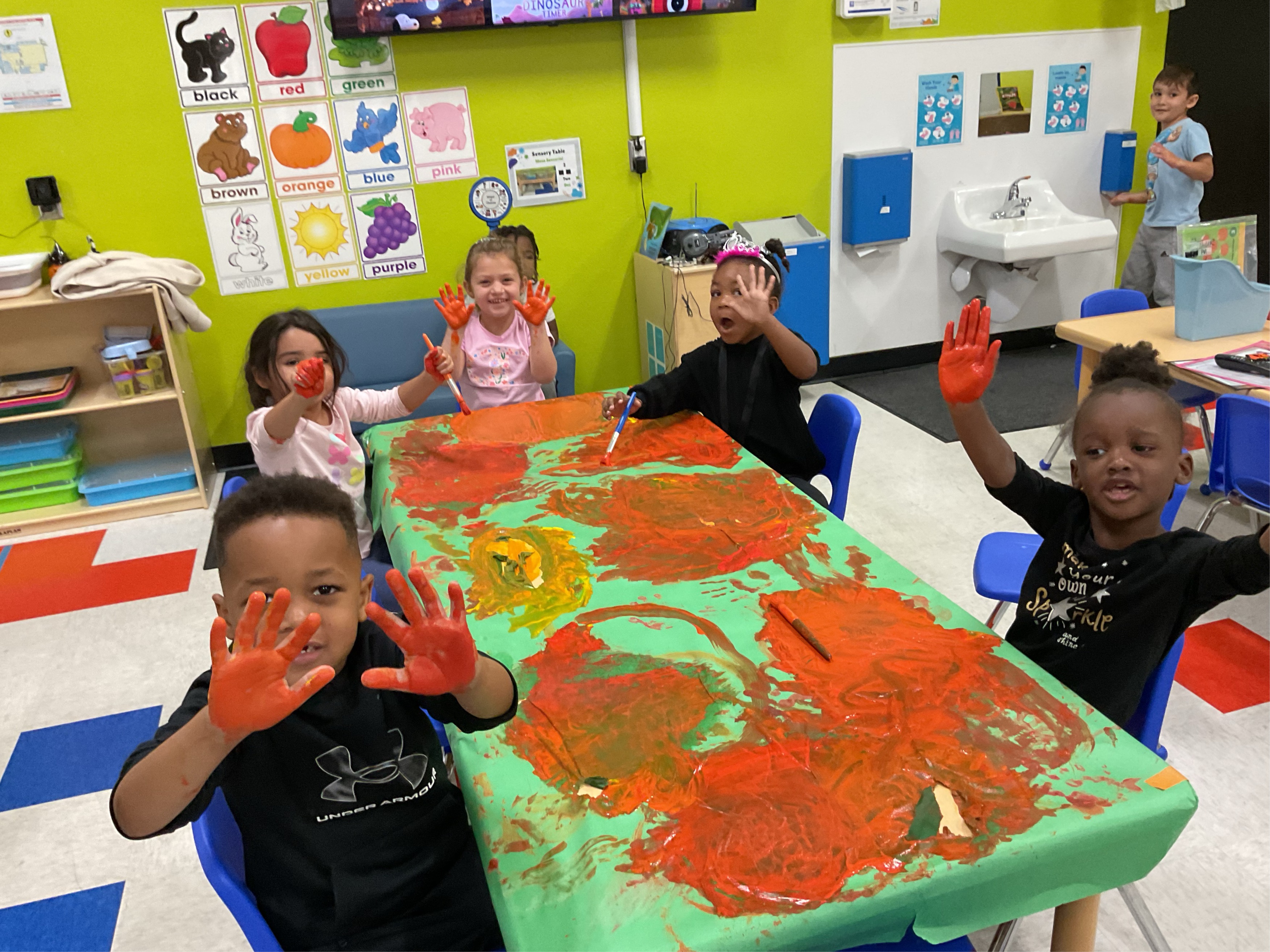 Classroom setting with 5 toddlers sitting around a table with orange paint all over it and their hands. Smiling for the photo. 