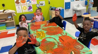 Classroom setting with 5 toddlers sitting around a table with orange paint all over it and their hands. Smiling for the photo. 