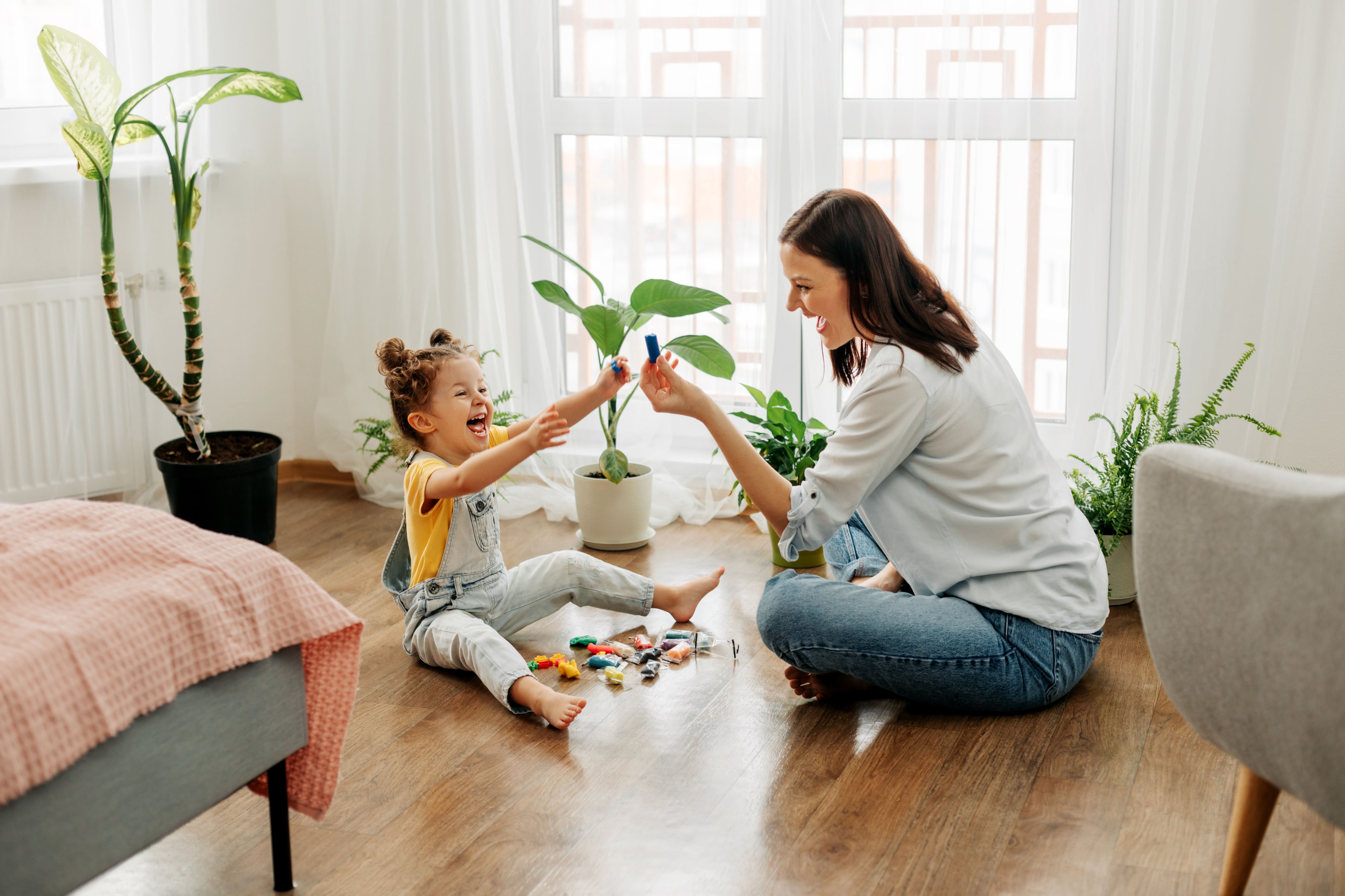 Young girl and women sitting on a hardwood floor in a living room setting with multiple green plants around, playing with toys, both laughing.