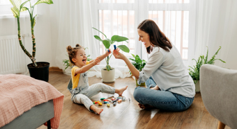 Young girl and women sitting on a hardwood floor in a living room setting with multiple green plants around, playing with toys, both laughing.