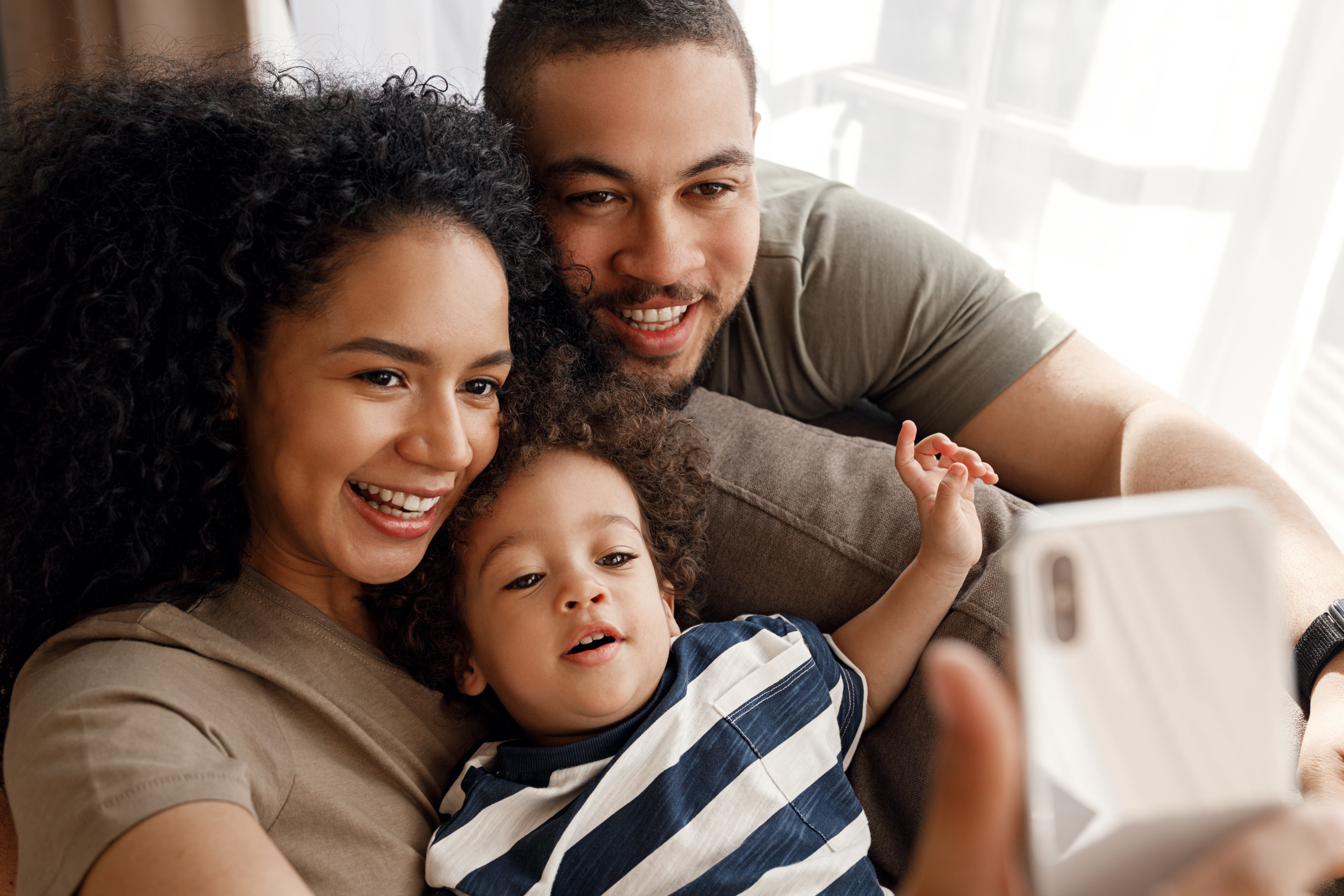 Adult female and male smile while taking a selfie with toddler in their lap wearing blue striped shirt.