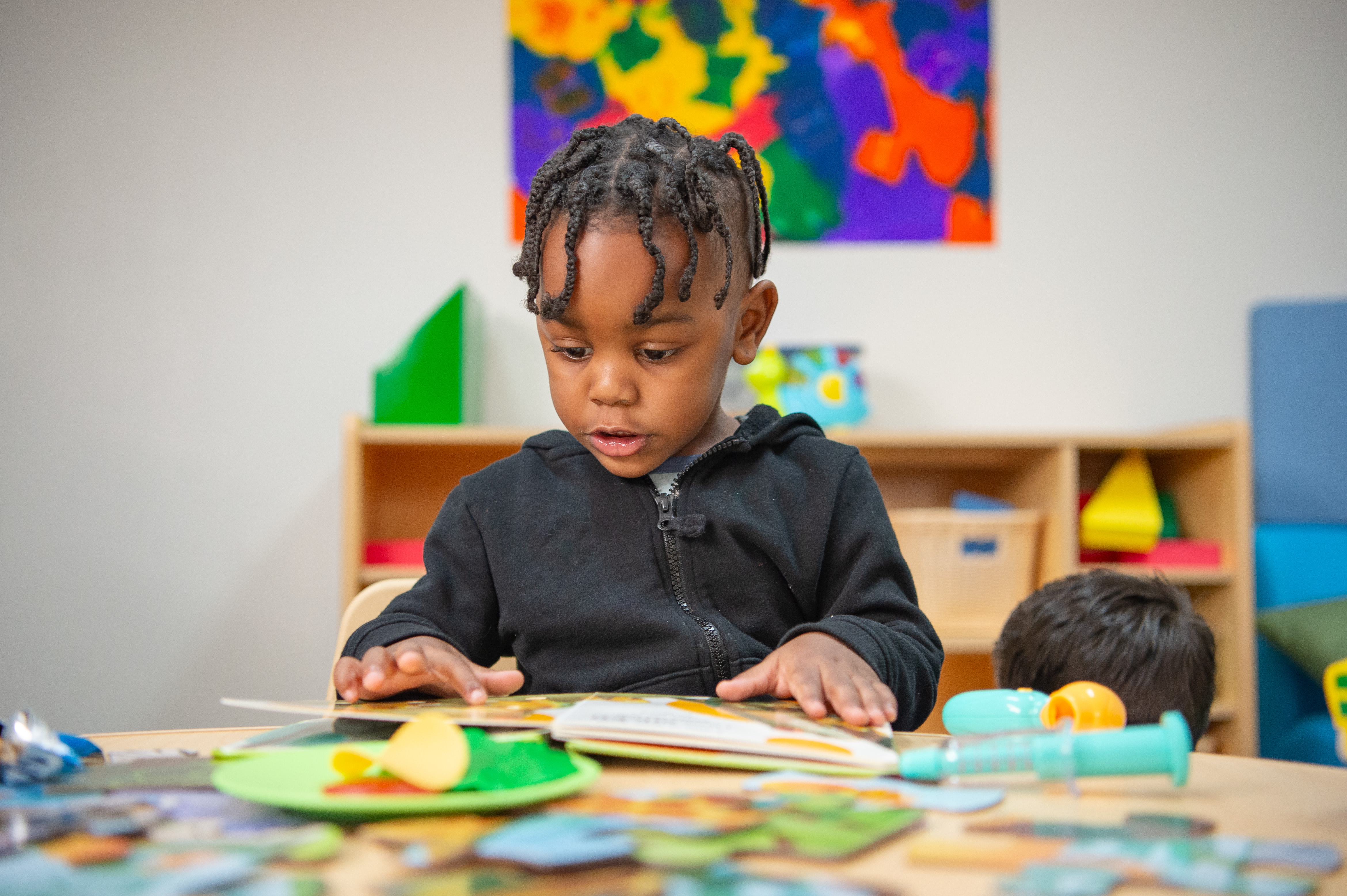 Little boy in a colorful classroom setting looks down and reads a children's book while sitting down.