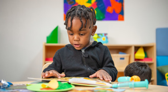 Little boy in a colorful classroom setting looks down and reads a children's book while sitting down.