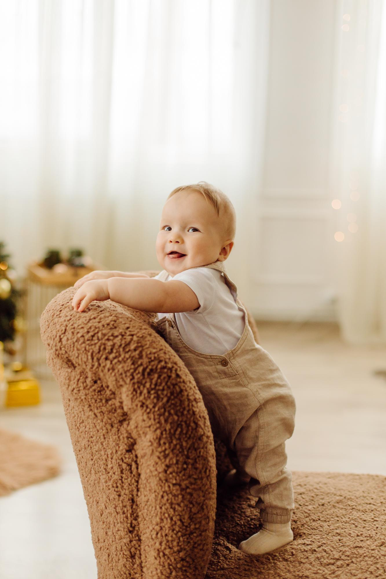 Toddler smiles while standing on a cozy looking fuzzy brown colored chair in a house setting. 